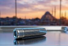 A photorealistic close-up of a sterile, titanium dental implant on a clean surface. In the heavily blurred background, the Istanbul, Turkey skyline is visible through a window during a warm sunset, representing dental tourism.