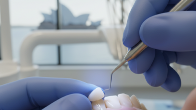 Close-up of a dental professional performing composite bonding on a patient's front tooth. Gloved hands are precisely shaping a tooth-colored resin material, with a subtle blue curing light visible. The background is a softly blurred, modern dental clinic, featuring a very faint and indistinct Australian city landmark through a window.