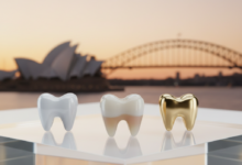 Close-up, ultra-detailed view of three distinct dental crowns made from different materials: a bright white all-ceramic crown, a slightly translucent zirconia crown, and a golden metallic alloy crown, all arranged on a reflective white surface. The background is a very soft, out-of-focus image of the Sydney Opera House or another iconic Australian landmark under a warm light. No text is visible.