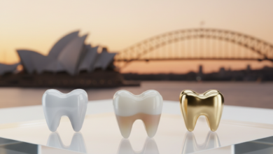 Close-up, ultra-detailed view of three distinct dental crowns made from different materials: a bright white all-ceramic crown, a slightly translucent zirconia crown, and a golden metallic alloy crown, all arranged on a reflective white surface. The background is a very soft, out-of-focus image of the Sydney Opera House or another iconic Australian landmark under a warm light. No text is visible.