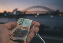 A close-up, photorealistic image of a hand holding blurred Australian dollar notes and coins, with a clean dental instrument in the foreground. In the soft-focus background, the iconic Sydney Opera House and Harbour Bridge are visible at dusk, symbolizing the financial burden of dental care in Australia.