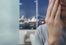 Close-up, photorealistic image of a person holding their jaw in discomfort, hinting at a dental emergency. The background is a blurred, iconic night view of the Sydney skyline, including the Opera House and Harbour Bridge. The scene uses soft, cool lighting and shallow depth of field.
