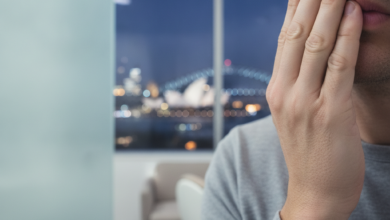 Close-up, photorealistic image of a person holding their jaw in discomfort, hinting at a dental emergency. The background is a blurred, iconic night view of the Sydney skyline, including the Opera House and Harbour Bridge. The scene uses soft, cool lighting and shallow depth of field.