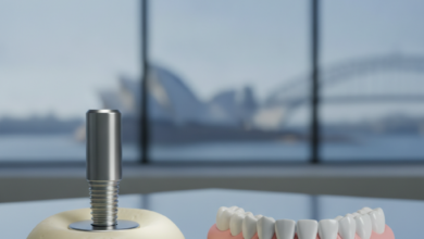Conceptual visual comparing a metallic dental implant next to a gum-colored acrylic denture, both displayed on a sterile surface in a modern dental clinic. A very blurred iconic Australian landmark is visible in the background through a window. The image is clean, professional, and focuses on the two distinct dental solutions.